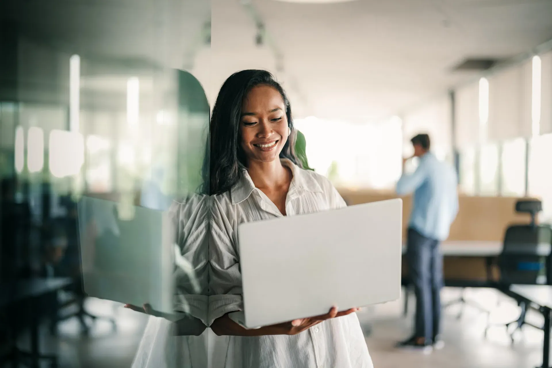 Smiling woman in a white blouse holding an open laptop in a bright modern office, with a colleague blurred behind her. Smiling woman in a white blouse holding an open laptop in a bright modern office, with a colleague blurred behind her.